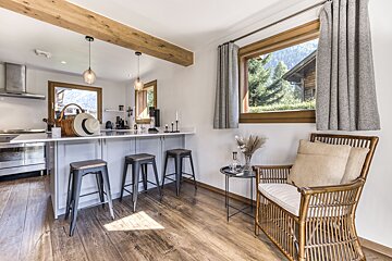 A bright, rustic-modern kitchen with a bar, stools, and exposed wooden beams. A wicker armchair by a window offers views of green trees and mountains.
