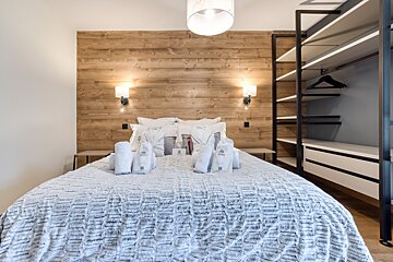 A modern bedroom features a large bed with white bedding, towels, and robes, set against a warm wood-paneled wall. Wall sconces provide light, and an open wardrobe is visible.
