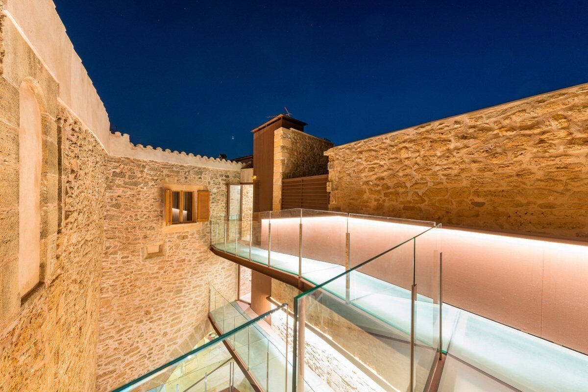 Sleek glass bridges with glowing lights connect rustic stone buildings at night, under a star-dusted dark blue sky.