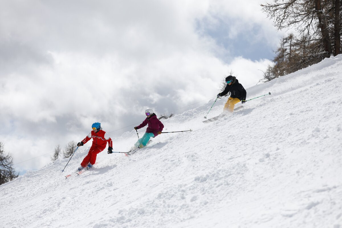 Three skiers in colorful gear carve down a snow-covered mountain slope under a cloudy sky with trees in the background.
