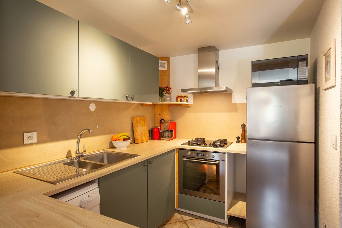 A cozy, modern kitchen with light wood countertops, sage green cabinets, stainless steel appliances, a sink, stove, and microwave. A fruit bowl adds color.