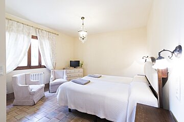 A bright bedroom with two single beds, two armchairs, a TV on a dresser, and a window with sheer curtains. The floor is terracotta tiled.