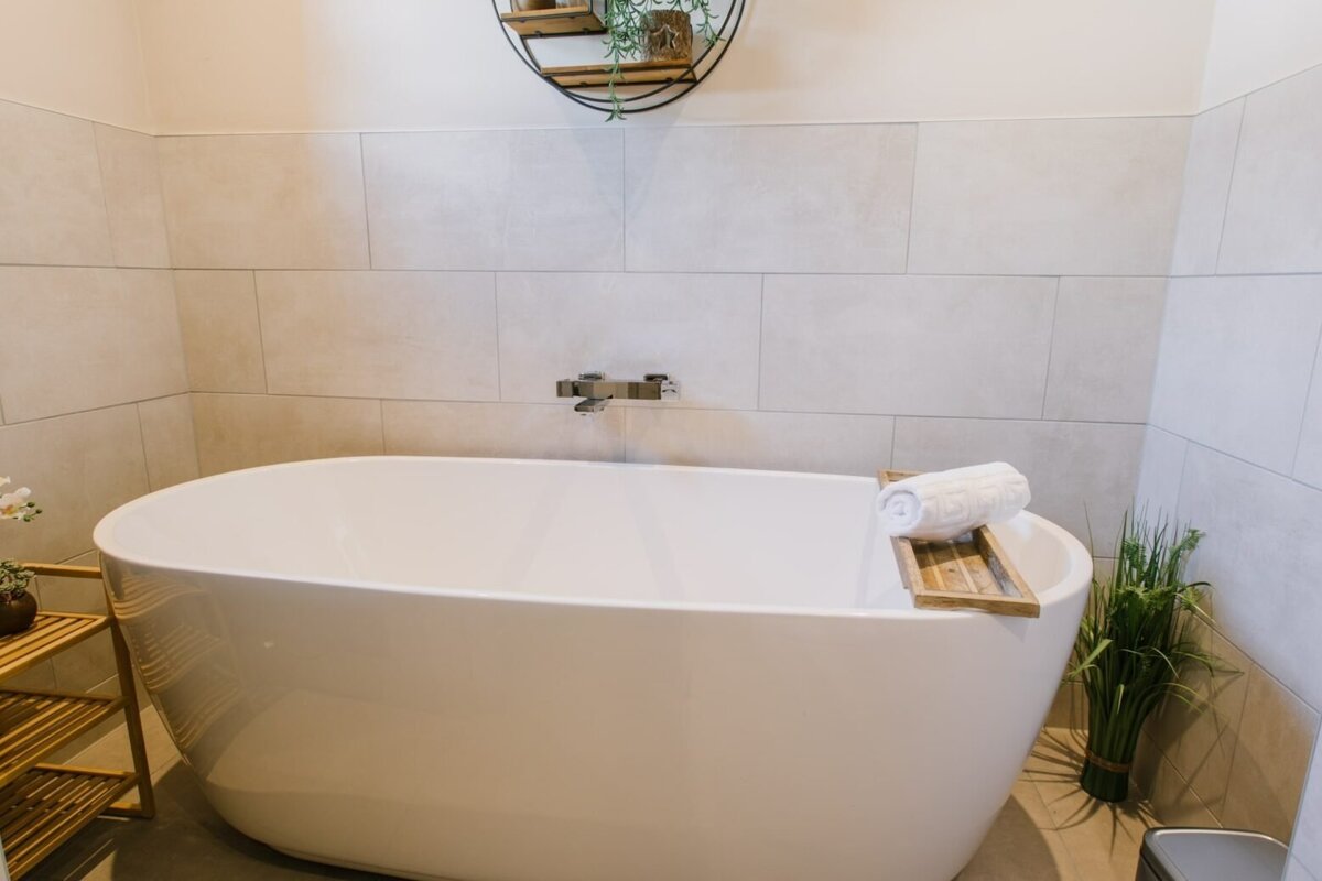 A contemporary bathroom features a white freestanding tub with a wooden caddy and towel, light-colored tiled walls, and decorative plants.