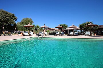 A beautiful turquoise swimming pool with straw umbrellas, lounge chairs, and palm trees under a bright blue sky, suggesting a relaxing resort.