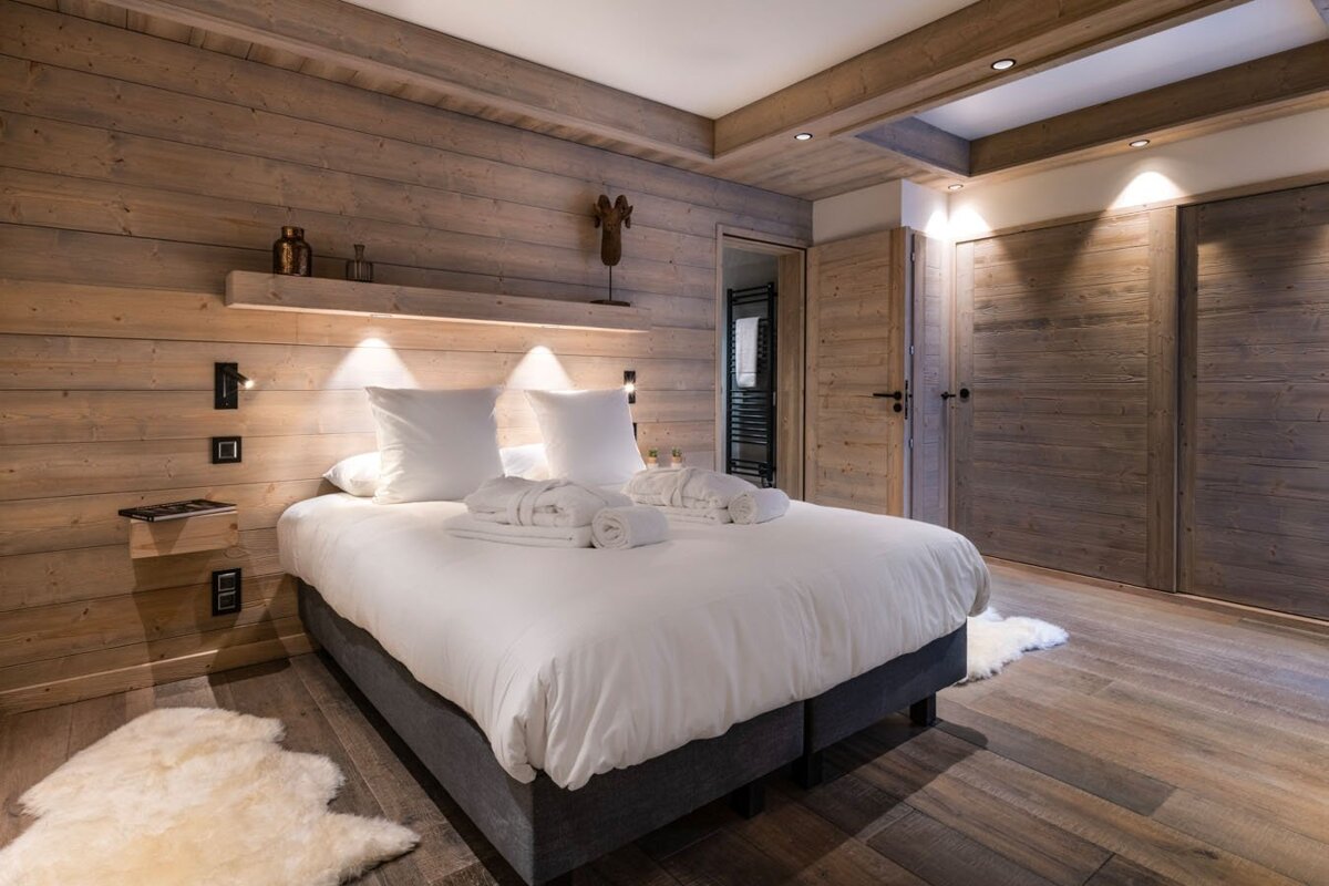 A cozy, wood-paneled bedroom features a large bed with white linens, pillows, and bathrobes. Soft lighting illuminates the wooden walls and floor with a sheepskin rug.