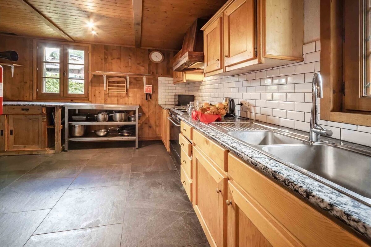A rustic wooden kitchen features white subway tile backsplash, granite countertops, a double sink, and kitchenware on shelves. A window shows greenery.