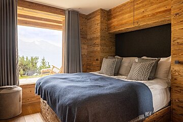 Rustic wood bedroom with a bed and blue blanket, featuring a large window showcasing a stunning mountain view.