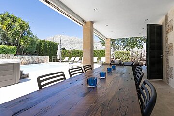 A spacious covered patio with a long wooden dining table, blue candles, and chairs, overlooking a pool, hot tub, and mountains.