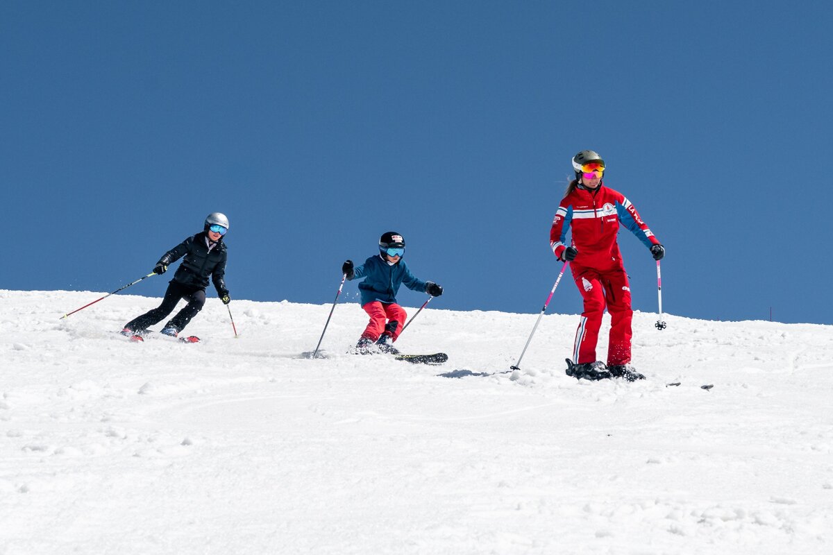 Three people, including a child, ski down a bright snowy mountain slope under a clear blue sky.