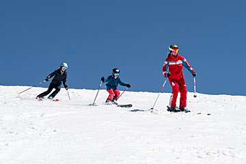 Three people, including a child, ski down a bright snowy mountain slope under a clear blue sky.