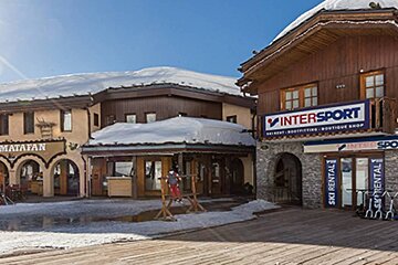 Sunny winter scene in a ski resort village with snow-covered chalet-style buildings, featuring an Intersport ski rental shop and a restaurant.