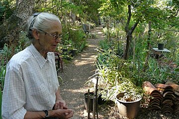 An elderly woman stands in a garden surrounded by potted plants