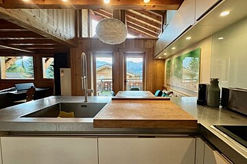 Rustic-modern kitchen with wooden beams, concrete counter, large sink, and expansive windows showing picturesque mountain chalets.