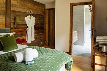 A chalet-style bedroom features rich wooden walls, a green quilted bed with fresh towels, and a white robe. A modern bathroom is visible through the open door.