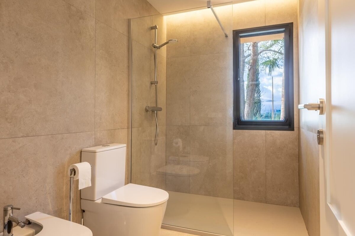 A modern bathroom with beige tiles features a toilet, bidet, and a glass shower, with a window offering a view of trees and water.