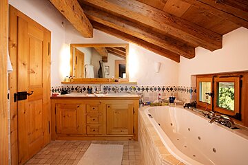 Rustic bathroom with exposed wood beams, double vanity, large tub, and a window. Tiled accents.