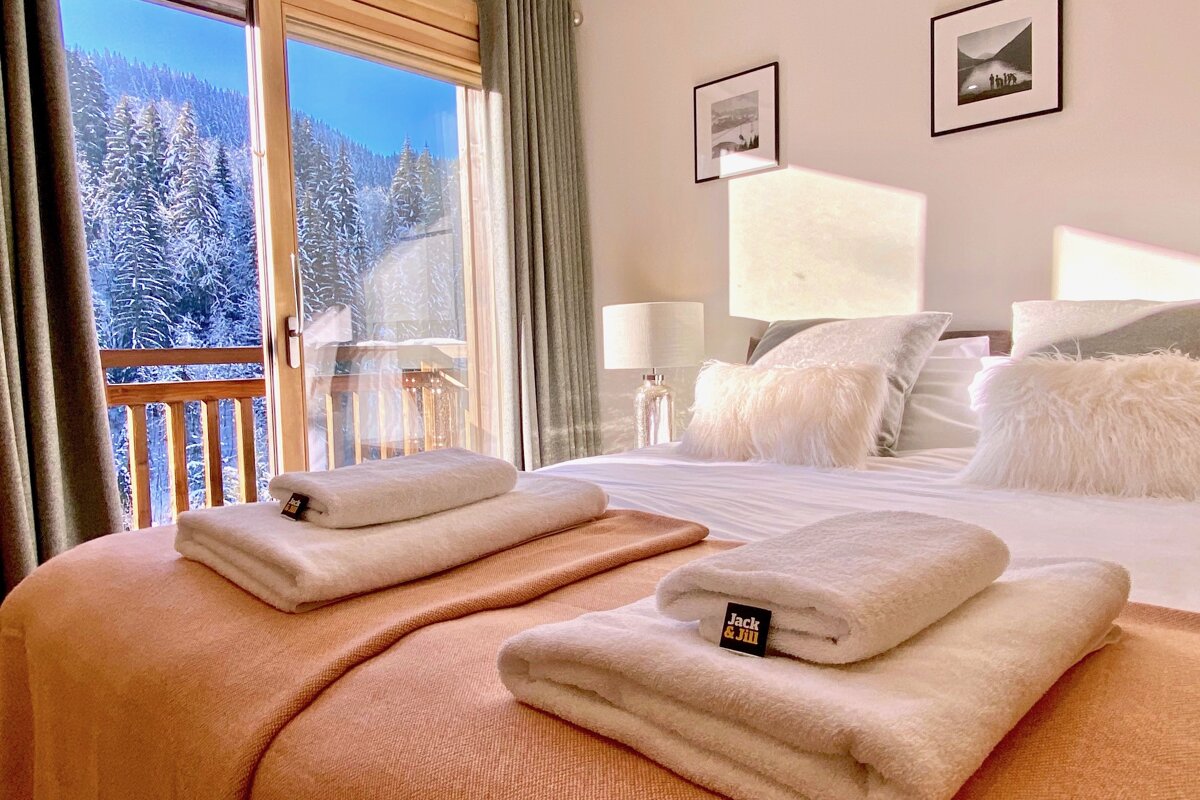 Sunlit bedroom with a cozy bed, white fluffy towels, and a stunning view of a snow-covered forest through a large window.