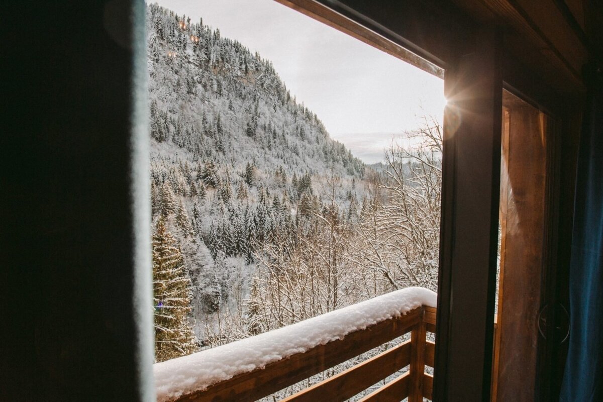 A sunlit, snow-covered mountain forest is viewed from a balcony with a snow-dusted wooden railing, capturing a serene winter scene.