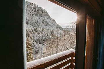 A sunlit, snow-covered mountain forest is viewed from a balcony with a snow-dusted wooden railing, capturing a serene winter scene.