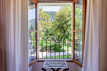 An open window with white curtains frames a lush green forest, distant hills, and a small balcony with a stool.
