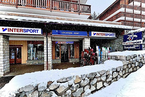 An Intersport ski rental shop in a snowy mountain town, with numerous skis lined up outside and a stone wall covered in fresh snow.