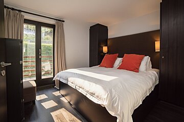A modern bedroom with a large bed, white bedding, orange pillows, and dark wood furniture. Glass doors open to a green view with sunbeams on the floor.