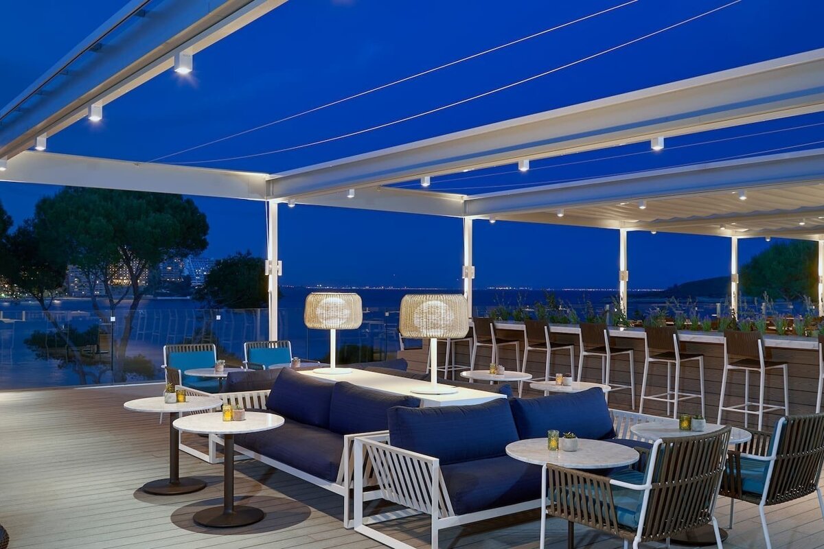 A chic outdoor lounge bar at dusk with modern blue and white furniture, overlooking a lit-up coastline and dark blue sea.