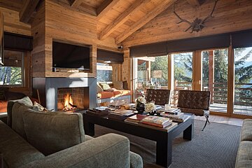 A luxurious, rustic ski chalet living room with a cozy fireplace, wood walls & ceiling, and panoramic mountain views through large windows.
