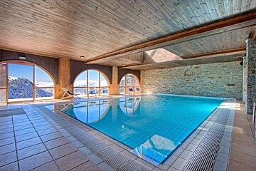 Indoor swimming pool with wooden ceilings, stone walls, and large arched windows overlooking snowy mountains.