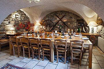 A rustic, arched stone dining room features a long wooden table set for dinner with warm lighting, candles, and traditional decor like a wagon wheel.