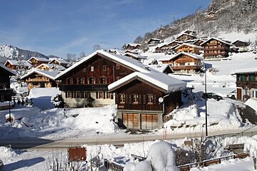 A charming alpine village with numerous wooden chalets blanketed in snow, nestled on a sunny mountain slope under a clear blue sky.