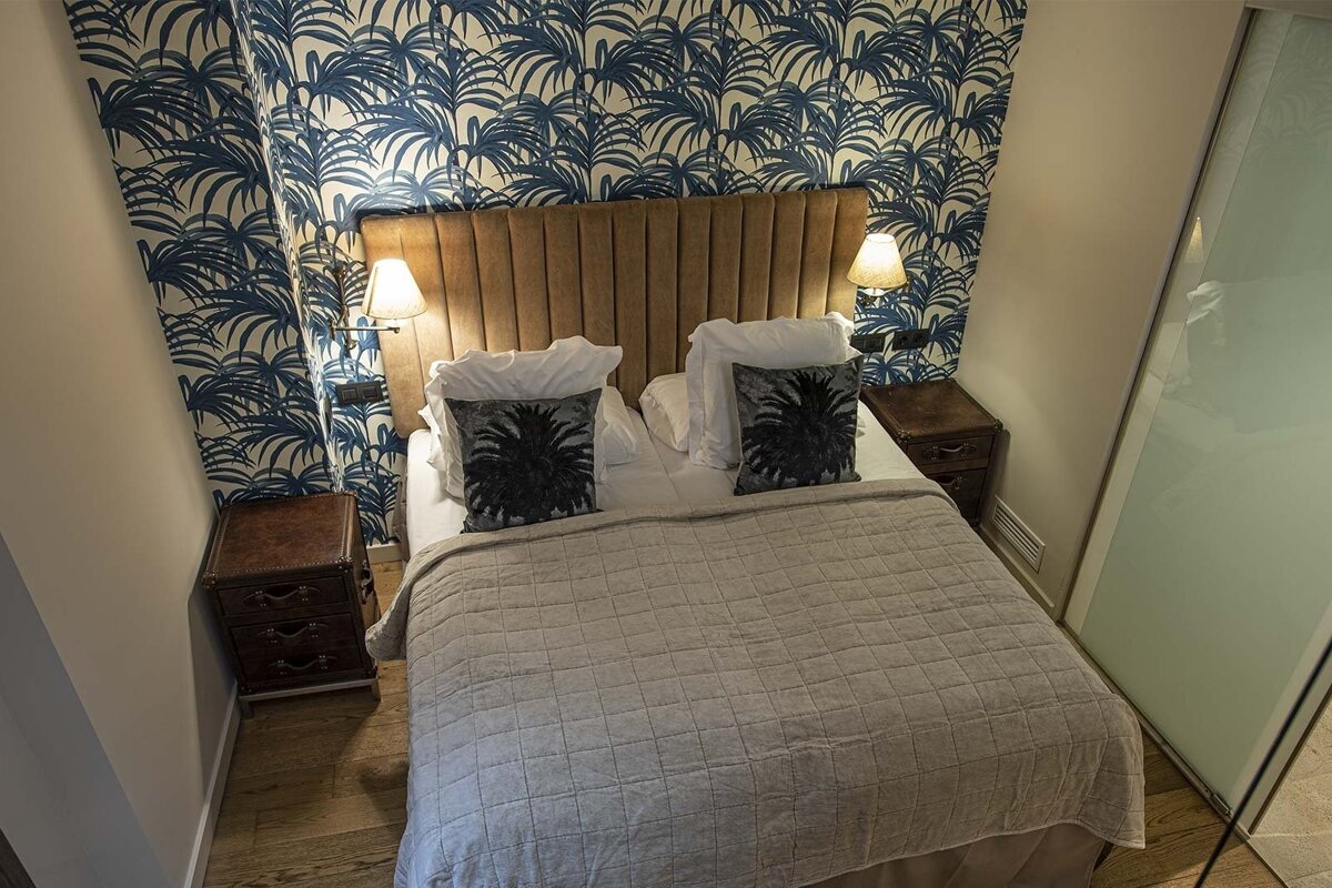 A cozy bedroom features blue palm leaf wallpaper, a padded light brown headboard, grey quilted bed, and dark bedside tables with lamps.