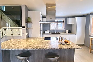 Bright, modern kitchen featuring a granite island with breakfast, bar stools, white cabinets, stainless steel appliances, and a glass staircase.