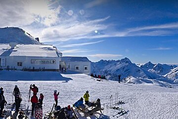 Skiers and snowboarders relax near a funicular station on a sunny, snow-covered mountain with majestic peaks in the distance.