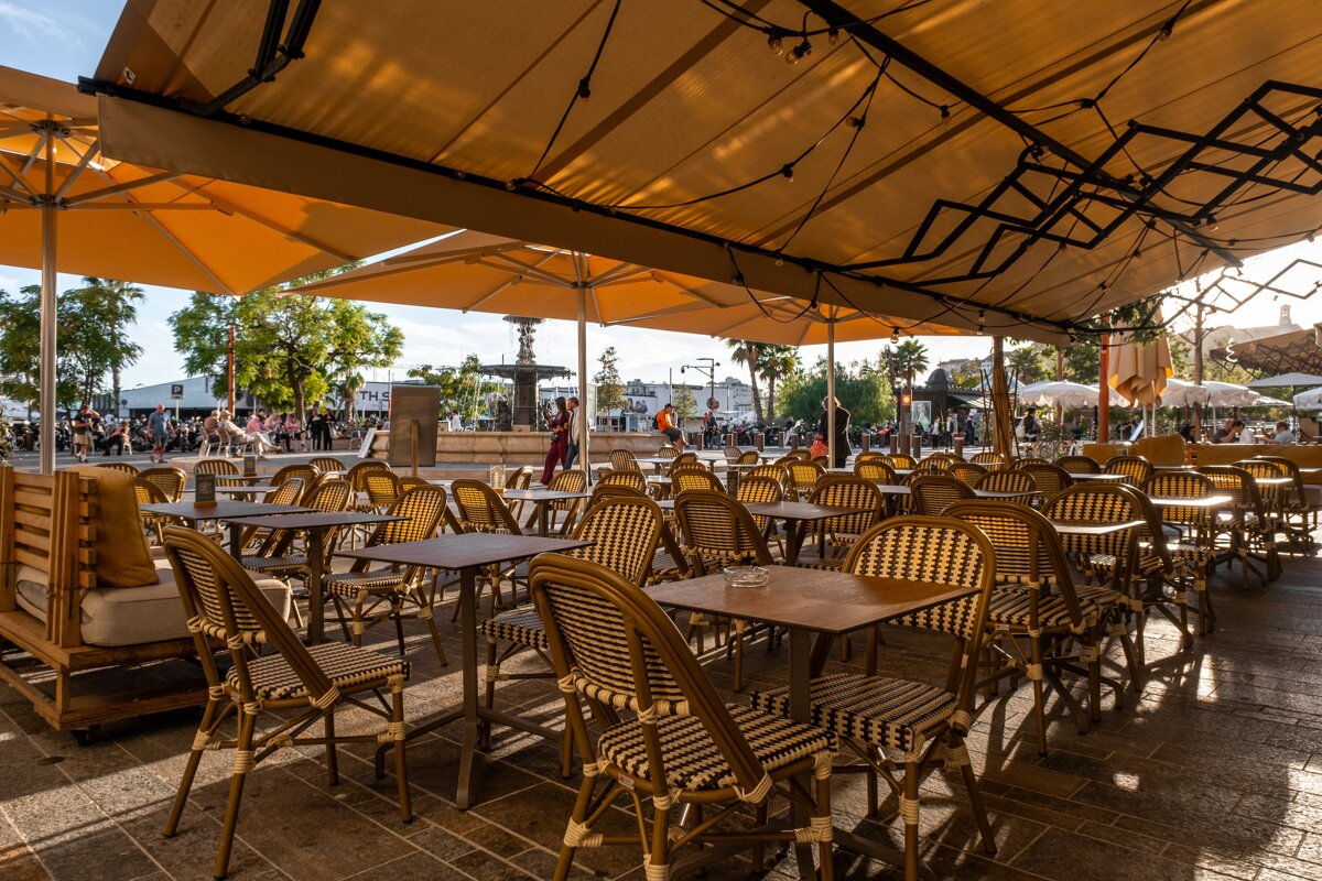 A sunny outdoor cafe with rows of empty rattan tables and chairs under large yellow awnings. A bustling square with people and a fountain is in the background.