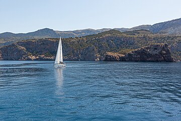 A white sailboat cruises on deep blue water, framed by rugged, forested mountains under a clear sky.