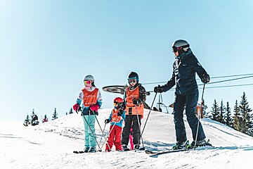 An adult and three children in ski gear, some with orange vests, stand on a sunny, snow-covered mountain slope under a clear blue sky.