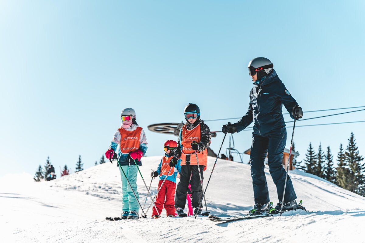 Four skiers, including three children in orange vests, stand on a sunny, snowy mountain with a clear blue sky and ski lift visible.