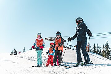 Four skiers, an adult and three children in orange bibs, stand on a sunny, snowy mountain slope under a clear blue sky with pine trees in the background.