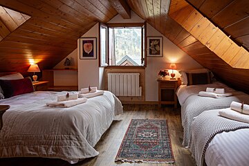 A cozy, wood-paneled attic bedroom features three beds with folded towels, soft lighting from lamps, a patterned rug, and a window with a scenic view.