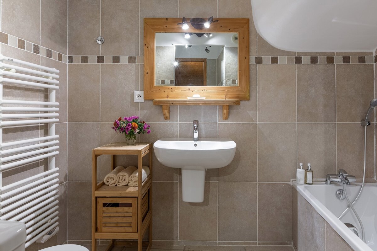 A beige-tiled bathroom with a wooden mirror over a white sink, a bathtub, a towel warmer, and a wooden shelf with rolled towels and flowers.