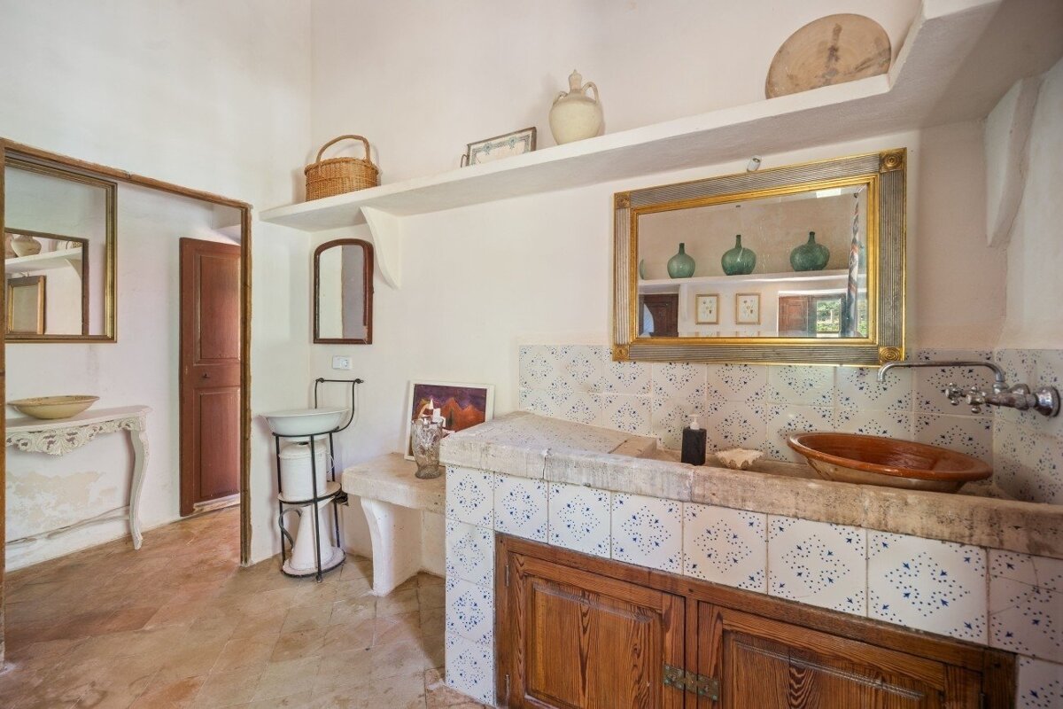 A rustic room with white walls, tiled counter with wooden cabinets, mirrors, shelves with decor, and multiple washbasins. Features a traditional, earthy style.