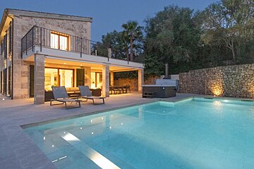 A luxurious stone house at dusk with an illuminated pool, hot tub, and outdoor lounge areas.