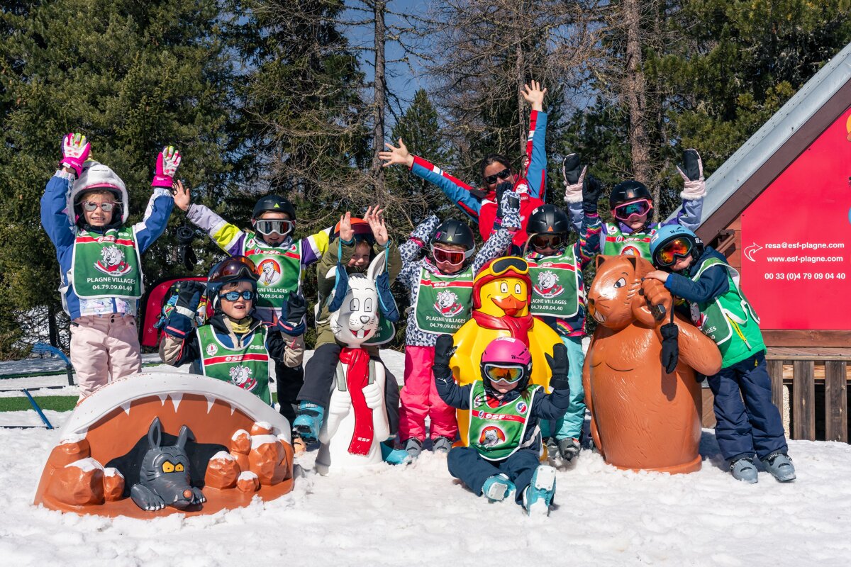 Cheerful ski school kids and an adult pose with fun animal mascots in the snow, hands up in excitement.