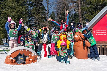 Happy ski school kids and an instructor pose with fun animal statues in the snow. They wear green bibs, showing excitement for skiing.