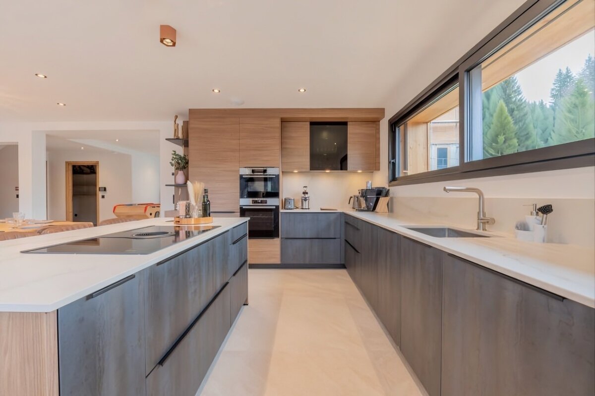 A chic, modern kitchen with a large island, two-toned wood and charcoal cabinets, integrated appliances, and a panoramic window framing a forest outside.