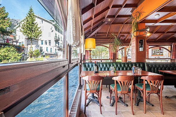 A cozy, rustic restaurant with large windows offering a direct view of a fast-flowing river, a white building, and lush trees outside.