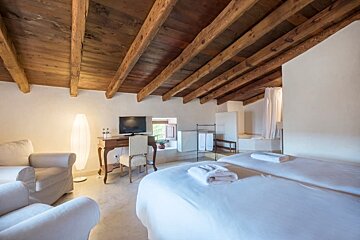 A bright, rustic room with an exposed wooden beam ceiling, two white beds, a desk with a TV, and a seating area with armchairs.