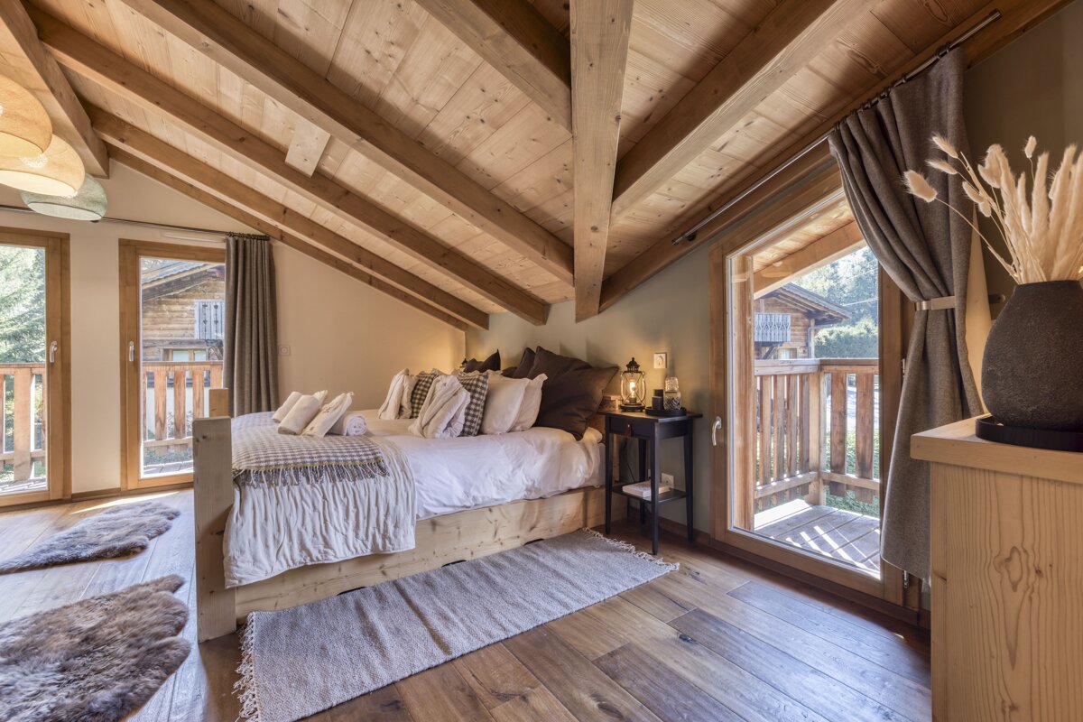 A cozy, rustic attic bedroom features a wooden vaulted ceiling, a large bed with pillows, and two balconies overlooking a chalet. Warm and inviting.