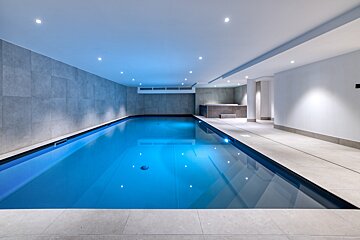 A modern indoor swimming pool with vibrant blue water, illuminated by ceiling spotlights. The room features grey tiled walls, light floors, and a small spa nook.